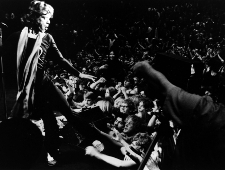 A man in a scarf on stage holds a microphone and dances for a large audience
