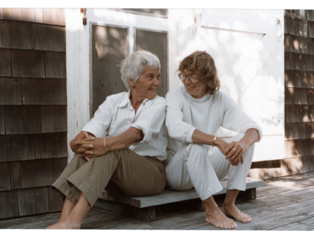 Two women sit on a doorstep smiling at each other. On the left is an older woman with short white hair in khaki pants and a white collared shirt. On the right is a woman with mid-length brown hair, glasses, a white turtleneck and white pants. Both are barefoot