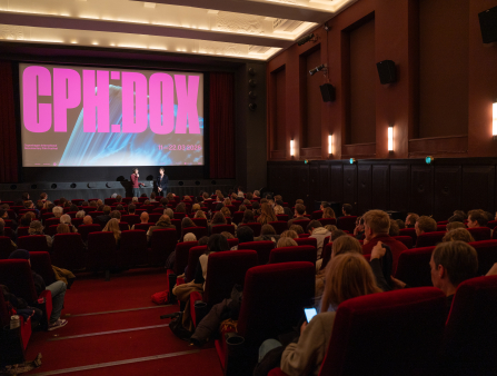 A read theatre full of people look at two figures on stage in front of a large screen which reads "CPH:DOX" in pink lettering