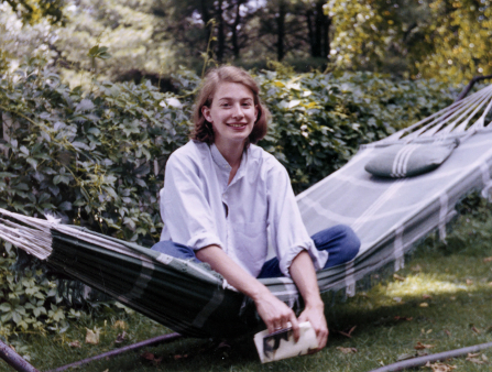 A young woman with a blue collared shirt and shoulder length brown hair sits in a grey hammock in front of an ivy covered fence. She holds a book and smiles for the camera