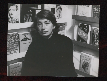 Black and white photo of a  young woman in a black shirt with shoulder length dark hair sitting in a corner. On both walls behind her are displays of books