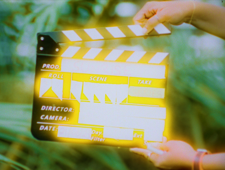 Two hands prepare to clap shut a slate in front of ferns. The text on the slate is obscured by shining yellow light