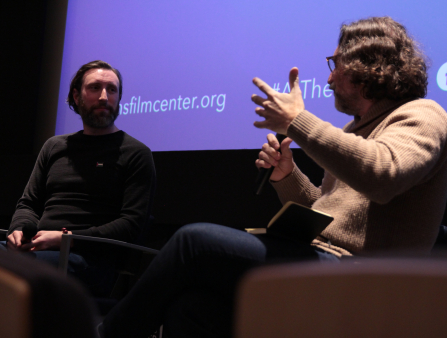 Two men sit in armchairs on stage in front of a large screen. On the left is a white man with beard and shoulder-length hair wearing a black sweater. On the right is a man gesturing his hand and holding a microphone, facing the man on the left. He wears a brown sweater and glasses, has shoulder length hair, and a notebook sits on his lap