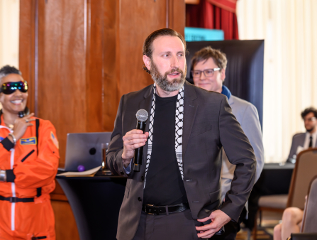 A man in a grey suit and black shirt with a beard (Alex Megaro) holds a microphone. Behind him and to the left is a woman in an orange space suit and sunglasses, and a man in a grey sweater with glasses. More people sit in the distant background