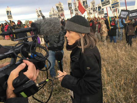 A middle-aged white woman in a black jacket and a grey wool hat stands in front of a protest holding a mic speaking into a camera