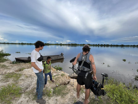 A man in a white T-shirt, jeans, and a black fanny pack and a man in a vest with a camera and microphone listen to a young boy in a green jersey. The three stand on the bank of a wide river