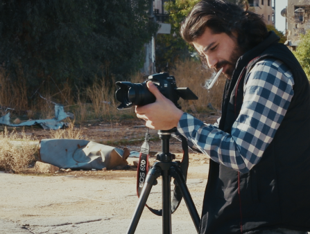 A man with long dark hair and a dark beard, wearing a black vest over a blue flannel shirt, operates a camera on a tripod while smoking a cigarette in front of a vacant lot