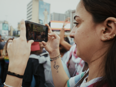 A woman photographs a chanting crowd with her phone held horizontally. She has a tattoo of a treble clef on her right wrist