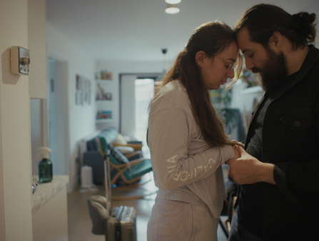 A woman with long dark hair in white and a man with a dark beard and long dark hair in black hold each others hands and touch foreheads in a white apartment