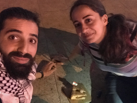 Selfie taken by a man with long dark hair and a dark beard, wearing a scarf around his neck, and a woman with long dark hair sitting on the ground