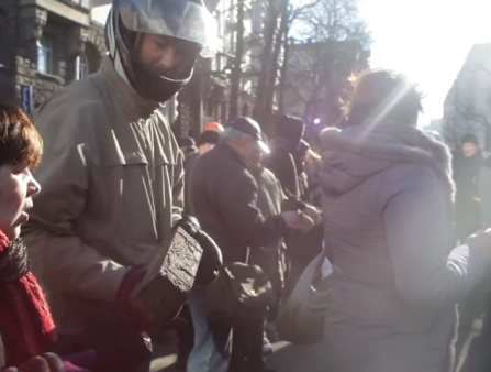 A man in a helmet hands an older woman in a red scarf a brick. They stand in a crowd, illuminated by bright sunlight