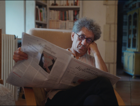 A man with grey curly hair and glasses sits in a white armchair and rests his head on his hand while reading the French newspaper diplomatique. He is inside a room with a bookshelf, window, and radiator