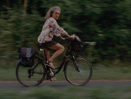 A woman with long grey hair, red glasses, a floral top, a short black skirt and sandals rides a black bike with a black bike bag along a forested path while looking to her right at the camera