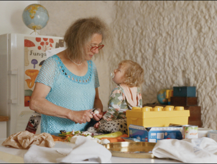 A woman with grey curly hair and glasses talks to a baby in a kitchen