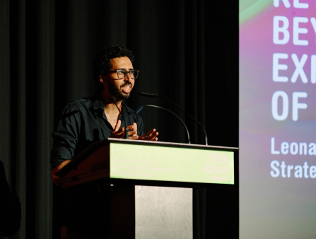 A man in a black shirt with black glasses and short dark hair and beard delivers a speech at a podium. Behind him is a black curtain and the lefthand side of a pink and green digital display showing text which cannot be read