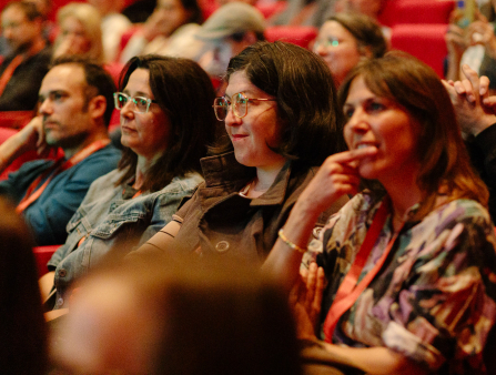 Three women and one man with orange lanyards sit in red theater seats, with more people blurry and partially visible in the rows behind and in front