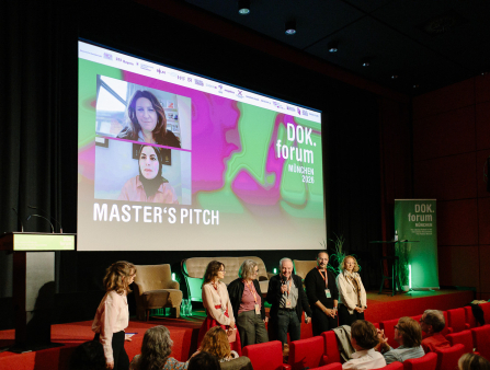 Two men and four women stand in front of a stage, wearing orange lanyards and facing a crowd. One man holds a microphone. On the stage behind them are two empty armchairs, an empty sofa, a vacant podium and a green banner which reads "DOK.forum MÜNCHEN" in addition to a large screen which displays the text "MASTER'S PITCH DOK.forum MÜNCHEN 2026" and two women who appear to be on video call
