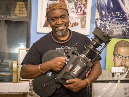 A smiling Black man in a brown hat, black T-shirt, and wooden and metal jewelry holds an old camera in front of awards and posters, one depicting Malcolm X
