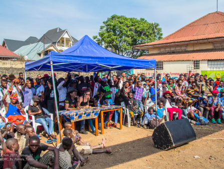 A large crowd of people sit in a clearing between houses and raise their fists. A blue tent in the center of the crowd covers a table behind which a man and a woman sit with pens and notebooks. A large speaker sits in front of them