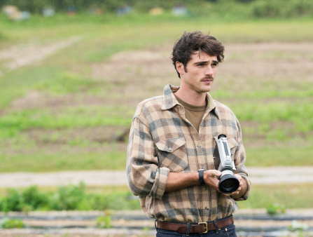 A young man with a mustache (Jacob Elordi) stands in a field, wearing a plaid shirt and holding a camera