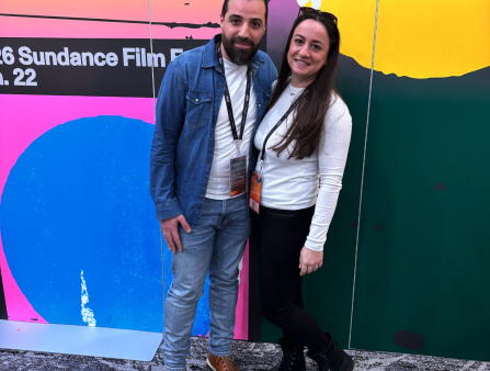A man with long dark hair and a dark beard, wearing a denim collared shirt over a white T-shirt, and a woman with long dark hair and sunglasses on her forehead, in a white shirt, stand in front of a colorful background where the words "2026 Sundance Film Festival Jan. 22" are written