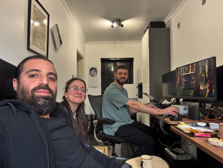Selfie of three people behind a desk. The man on the left who is taking the photo has a dark beard and long dark hair. The woman in the center has glasses and smiles. The man behind the desk operates a large computer and wears a blue shirt. He has short dark hair and a short beard