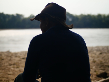 Shot from behind a man in a dark shirt and brown brimmed hat sitting on a riverbank staring across the water