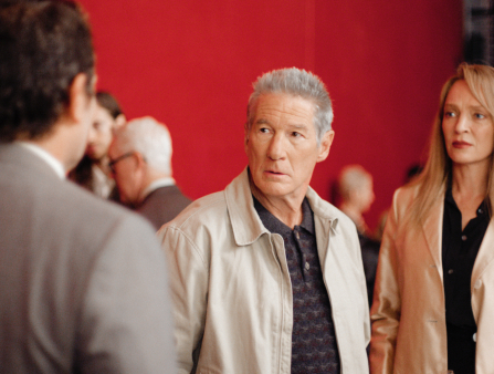 An older man in a white jacket with grey hair (Richard Gere) looks shocked at a man who faces towards him and away from the camera. A blonde woman to his right looks at him in apprehension. Behind them is a vivid red wall and blurry people mingling in the background