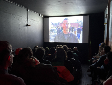 A theatre full of people look at a screen which displays a smiling man with a shaved head and green jacket