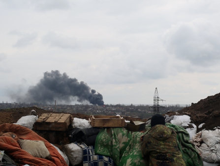 A soldier looks out from behind a rampart constructed of dirt and rubbish over a city from which a large plume of smoke rises
