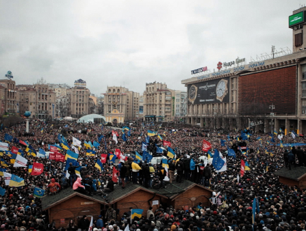 A massive crowd gathers in an open square, surrounded by large buildings. Many hold Ukrainian flags, some hold red and white flags and one holds a European Union flag