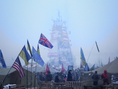 A tower rises through the fog. In the background is a building with columns. In the foreground is a crowd of people, large tents, and Ukrainian, British, American, and European Union flags