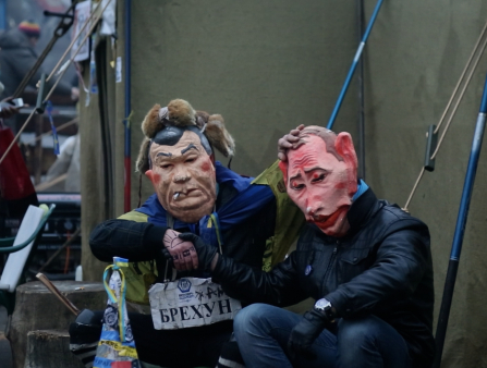 Two people in jackets and large masks sit. The masks are charicatures, one is pink with large ears and little hair, another has black hair and rosy cheeks and holds a cigarette in its mouth