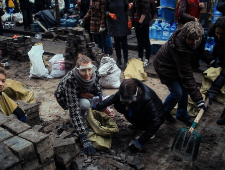 Two women in safety goggles look up at the camera in the midst of some kind of physical labor. They are surrounded by bricks and other people, two of whom hold the same shovel and one of whom squints at the camera. All are warmly dressed. Jugs of water sit in the background