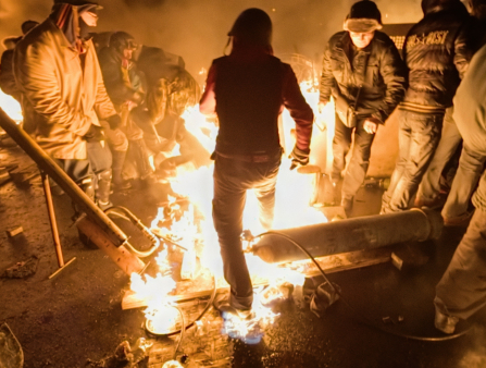 A group of men surround an open flame. One man in a helmet appears to attempt to stomp the fire out. All are warmly dressed while more fire and smoke fills the background