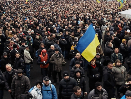 A massive crowd of people in winter clothes is gathered. One man towards the front waves a large Ukrainian flag