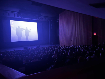 A theatre of people lit in purple stare at a screen displaying two soldiers pointing guns off-screen shrouded in mist