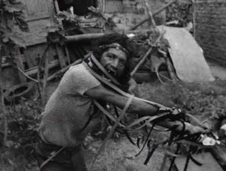 Black and white photo of a  man in some kind of scrapyard wrapped in straps of some kind staring wild eyed at the camera. He has long hair and wear a short-sleeved white shirt.