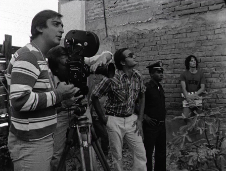 Black and white photo of five men in front of a brick wall, all observing something off screen to the right. On the left is a man in a striped shirt. Next to him a man operates a large, old camera on a tripod, and is obscured behind the machine. In the center is a man in a plaid shirt and sunglasses looking up. Next is a man in a police uniform. The man on the far right holds a slate and leans against the brick wall