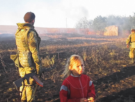 Two soldiers look away from the camera over a large scorched field. The nearer soldier holds a rifle. A little girl in a red jacket with blonde hair, nearest to the camera, sobs