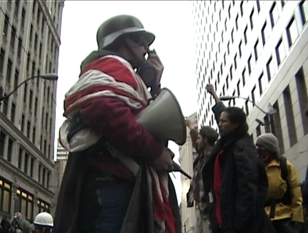 A man in an army helmet, wrapped in an American flag, holds a megaphone under his right arm and speaks into something he's holding in his left hand. He stands in between two tall buildings and three people behind him raise their arms to the sky