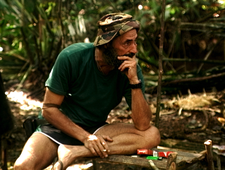 A man in a green T-shirt, shorts, and camo bucket hat sits with his head in his left hand, seemingly deep in thought. Two packs of cigarettes and a lighter sit on a wooden bench beside him. Jungle foliage fills the background