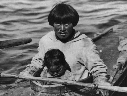 Black and white photo of a man with a bowl cut and a child in his lap sitting in a canoe