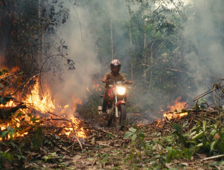 A man in a red motorcycle helmet on a red motorcycle with its headlight on drives through the center of a burning forest