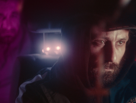 A bearded man looks grimly down while driving. Another cars headlights can be seen behind him. He wears a cap, hood, and jacket. Eerie, purple lighting