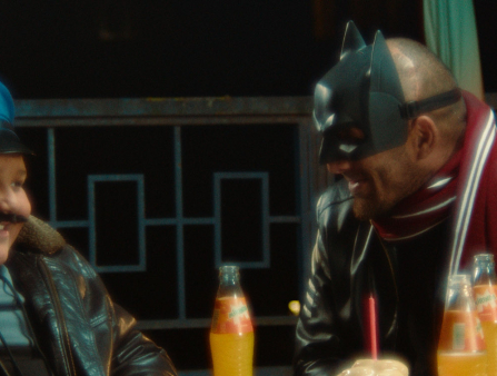 A young boy in a cop costume and a man in a batman mask laugh at each other. They sit at a table on which rests three orange sodas