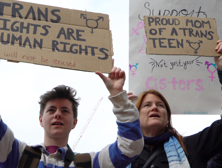 A young man and his mother hold up signs at a protest that read: "Trans Rights Are Human Rights" and "Proud Mom of a Trans Teen" written in cardboard box material.