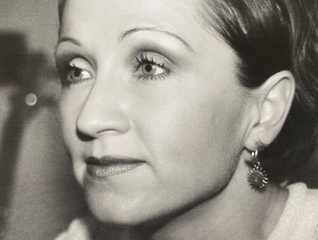 A black and white close-up shot of a woman's face. She has long dark hair, tied back, and dangly earrings