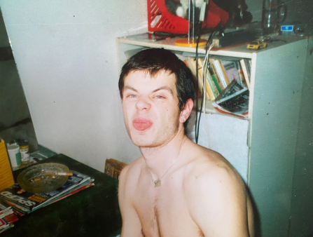 A young man with a necklace and no short sticks his tongue out at the camera. Shot with bright flash