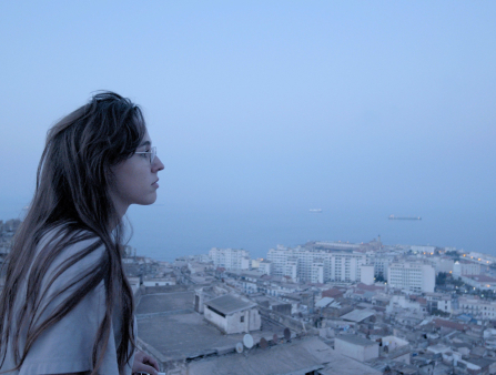 A woman in glasses with long brown hair looks over a city on the ocean at dusk